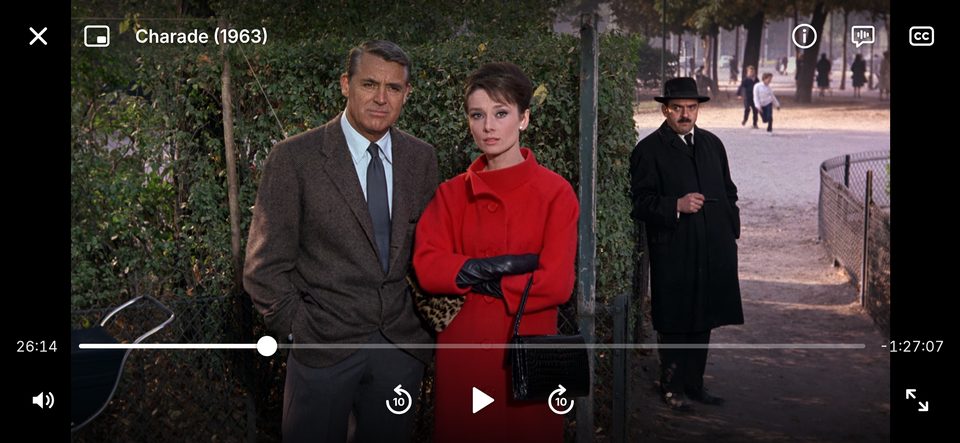 The Hi, bye player full screen on an iPhone in landscape, paused on Charade (1963) at 26:14: a man in a tweed jacket and a woman in a red coat stand by a park hedge while a man in a black coat and hat watches from the path.